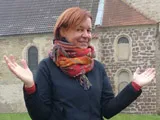 Eine Frau mit ausgebreiteten Armen vor einer Bruchsteinkirche